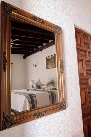 Un miroir doté d’un cadre en bois décoratif, reflétant une pièce où l’on aperçoit un lit, des oreillers et un luminaire fixé au mur.