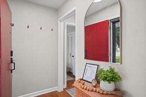 A hallway with a white door, a red door, and a mirror on the wall. There is a framed sign on a wooden table. - Ginger Sol - 2BR/1BA - Centrally Located (Lubbock)