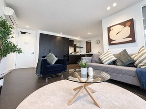 A modern living room with a glass coffee table, dark blue sofa, and a grey sofa with striped pillows. A kitchen is visible in the background. - Southbank Stunner with 2 Carparks and Dream Views (South Melbourne)