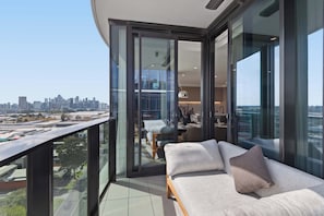 A modern balcony with a city view, a sofa with cushions, and a dining area visible through glass doors. - Jaw-dropping 4BD Penthouse in Beachside Resort (Port Melbourne)
