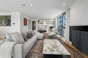 A modern living room with a sofa, coffee table, and a TV. There is a bookshelf and a chair in the background. - Jaw-dropping 4BD Penthouse in Beachside Resort (Port Melbourne)