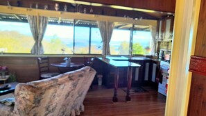 A room with a wooden floor, a sofa, a table, and a view through large windows. - Clives Manor is a cosy cabin nestled in the hills of Turondale. (Turondale)