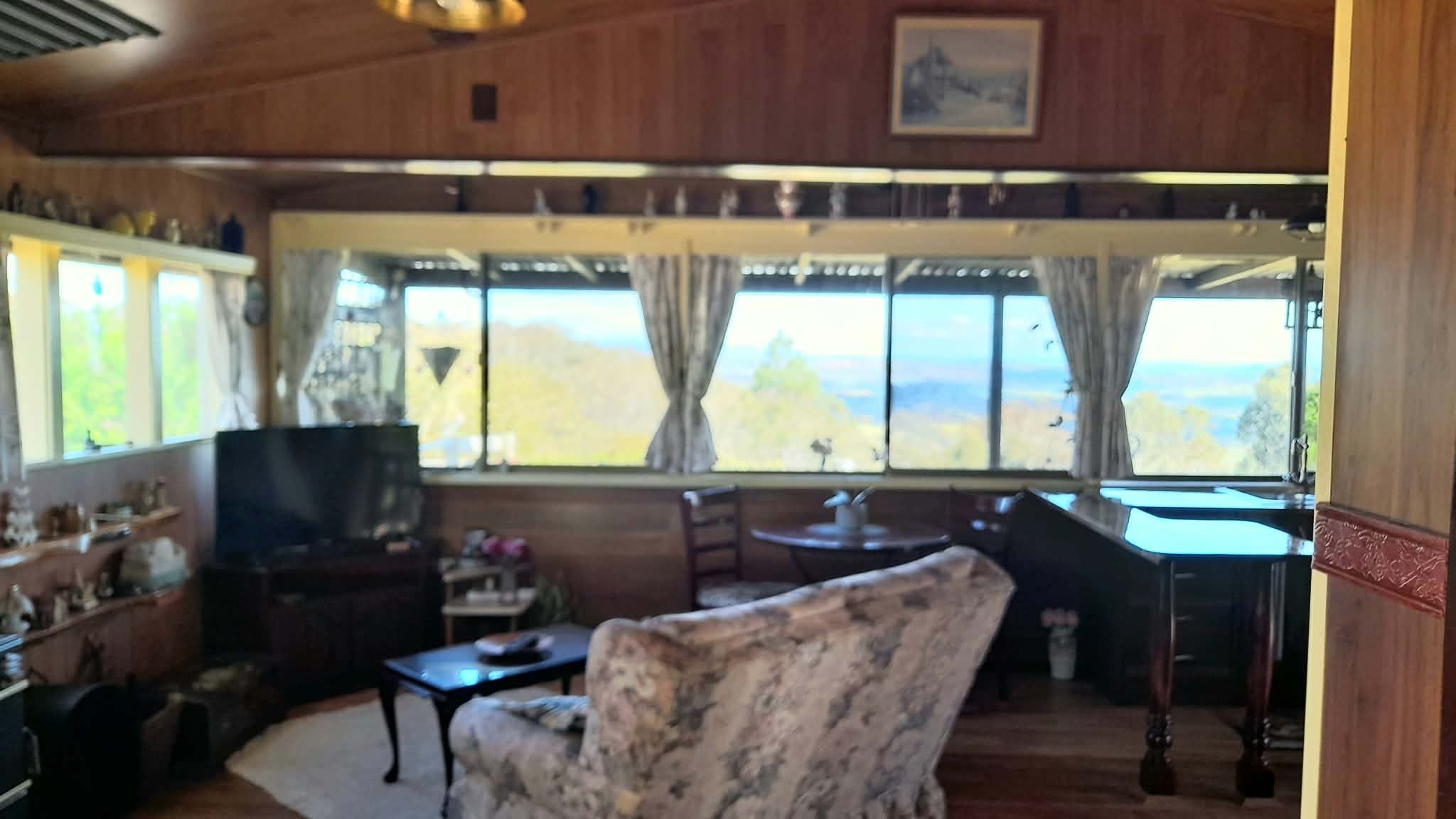 A living room with a television, a patterned armchair, a small table, and a view through large windows.