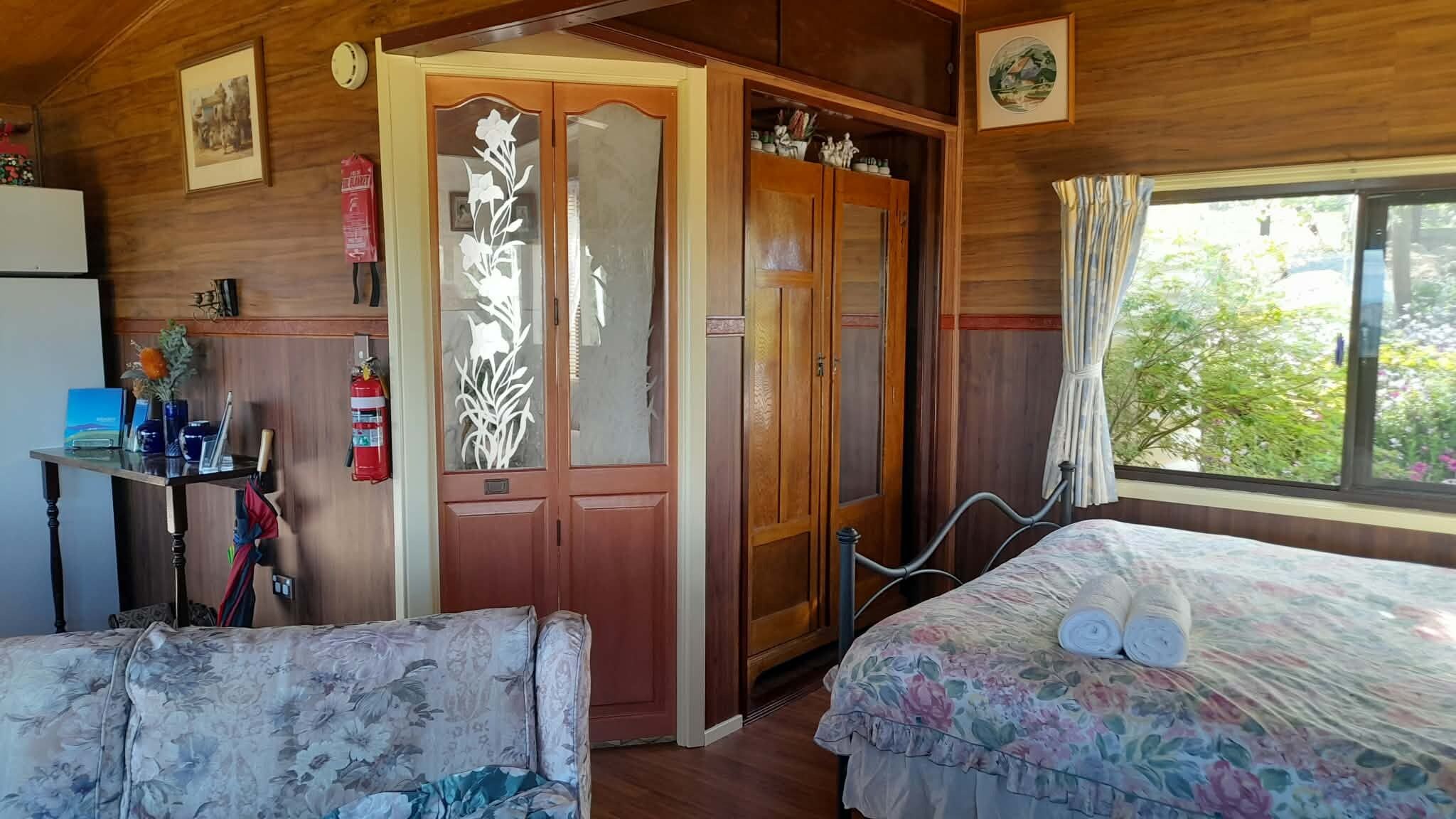 A room with two beds, a wooden wardrobe, a small table with a vase, and a window with a view of greenery.