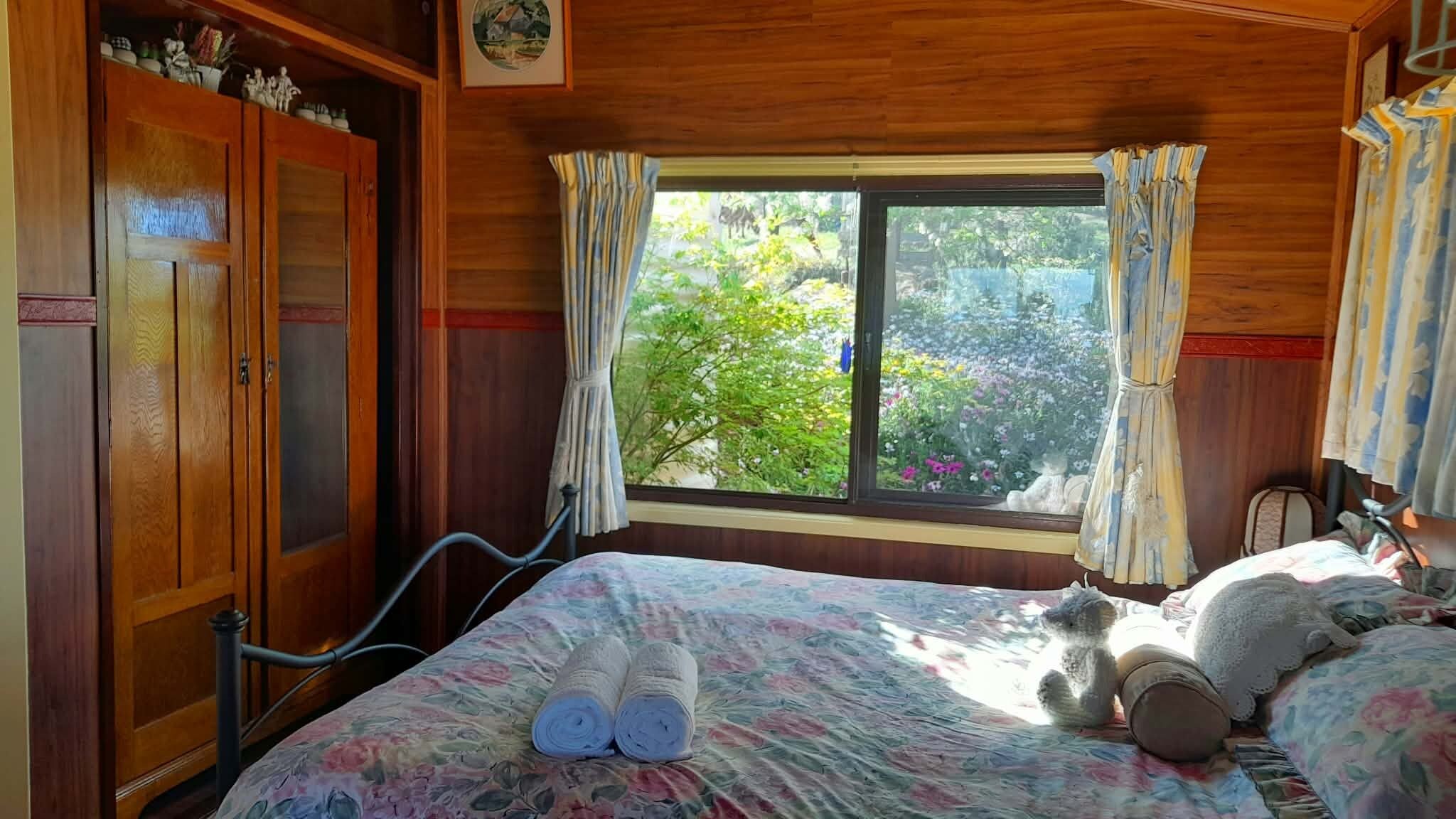 Ein Schlafzimmer mit einem Holzschrank, einem Bett mit Blumenmuster und einem Fenster mit Vorhängen, mit Blick auf Grünfläche.