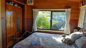 Ein Schlafzimmer mit einem Holzschrank, einem Bett mit Blumenmuster und einem Fenster mit Vorhängen, mit Blick auf Grünfläche.