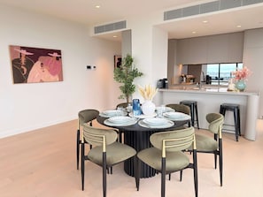 A modern dining area with a round table set for six, surrounded by upholstered chairs with gold accents. A potted plant and decorative vase are on the table. - Jaw-dropping Views from Brand New 3bd Luxe Home (Melbourne)