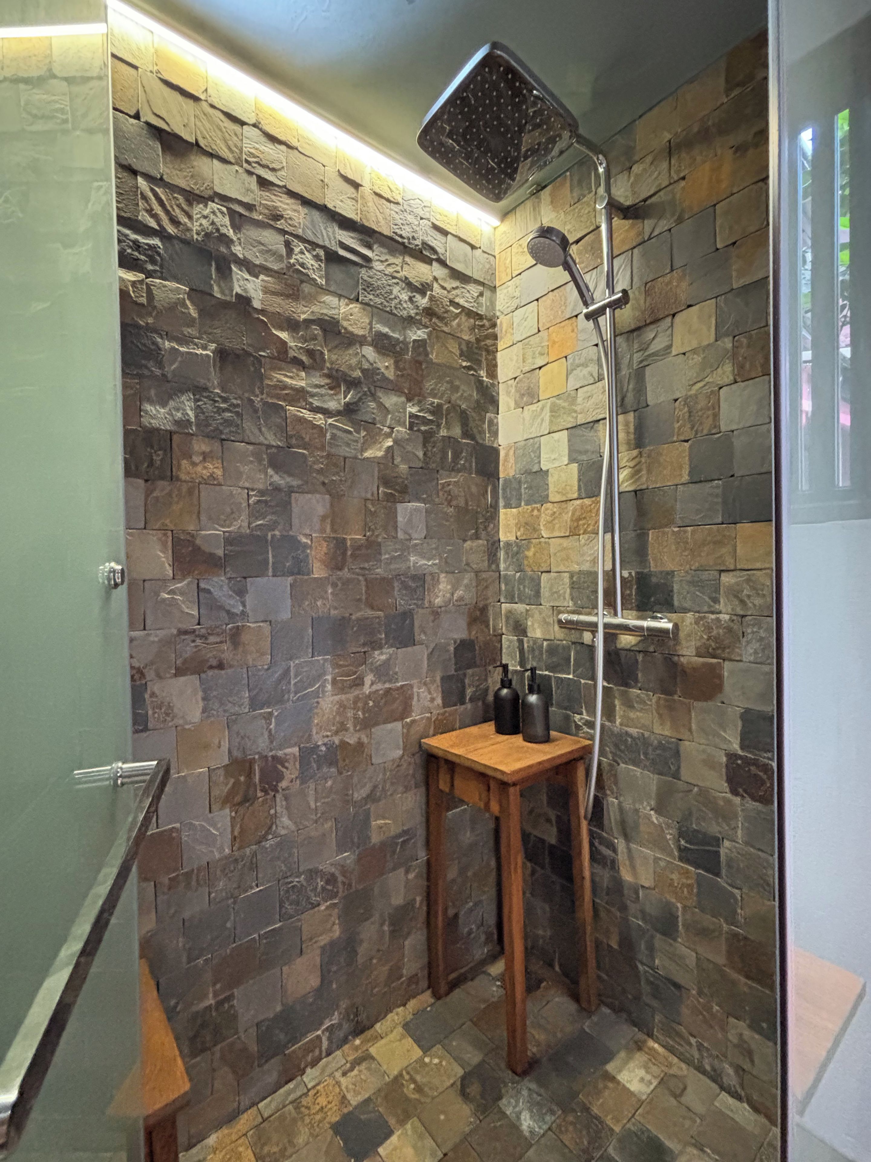 Eine Dusche mit Steinwand, einem hölzernen Hocker und einer Glastür.