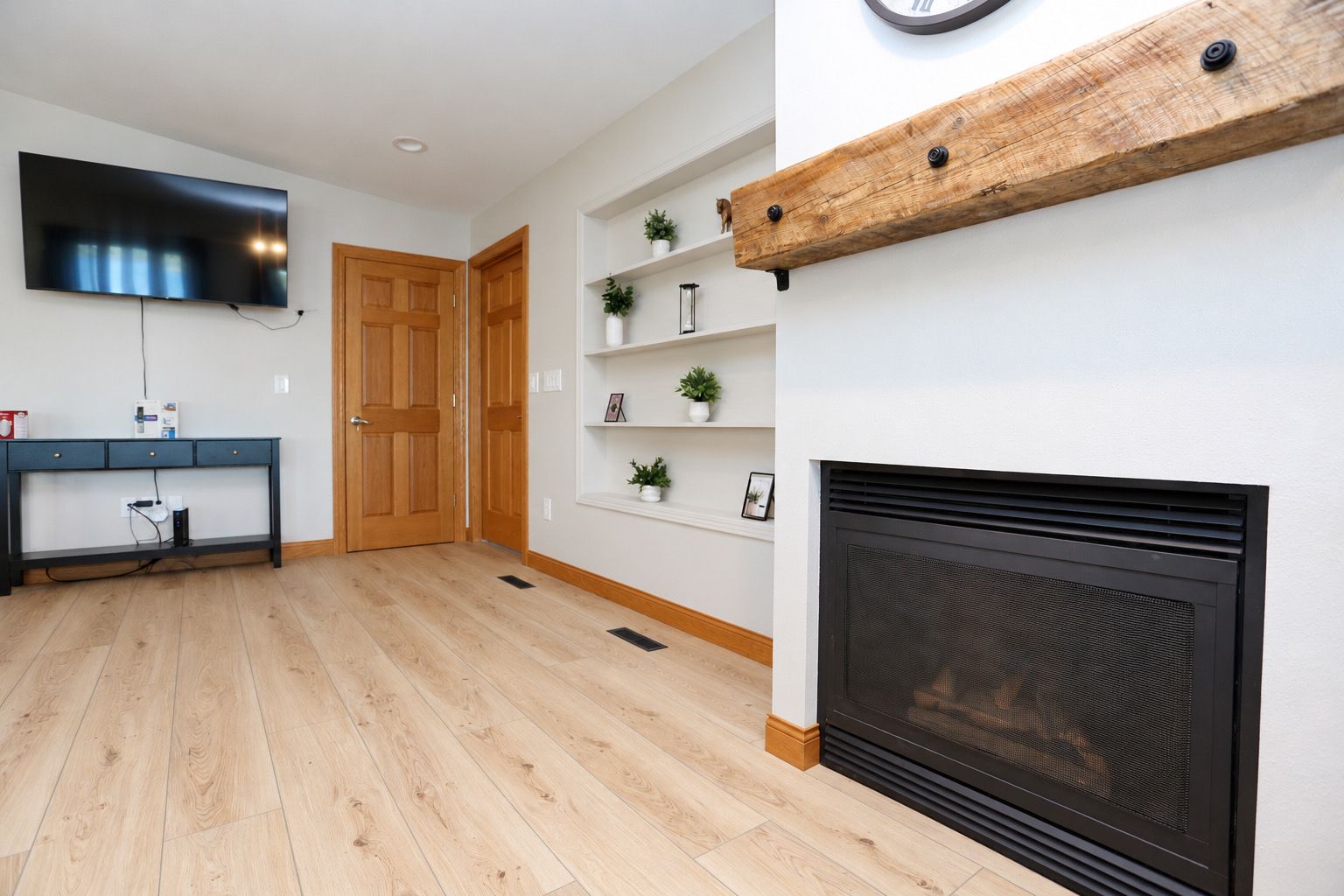 A modern living room with a flat-screen TV, a wooden door, a fireplace, and a wall-mounted shelf with decorative items.