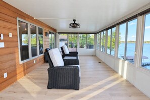 A covered outdoor area with wicker furniture, large windows, and a view of the water.
