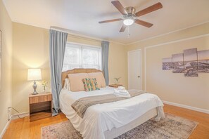 Un dormitorio con cama, mesita de noche, lámpara, ventilador de techo y ventana con cortinas.