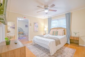 Un dormitorio con cama, ventilador de techo, mesita de noche, lámpara, ventana con cortinas y puerta que da a otra habitación.