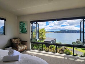 A bedroom with a large window, a bed, a chair, and a painting. - Hot Water Beach Panorama - Hot tub, sea views, 3 min walk to beach, pets allowed (Hot Water Beach)