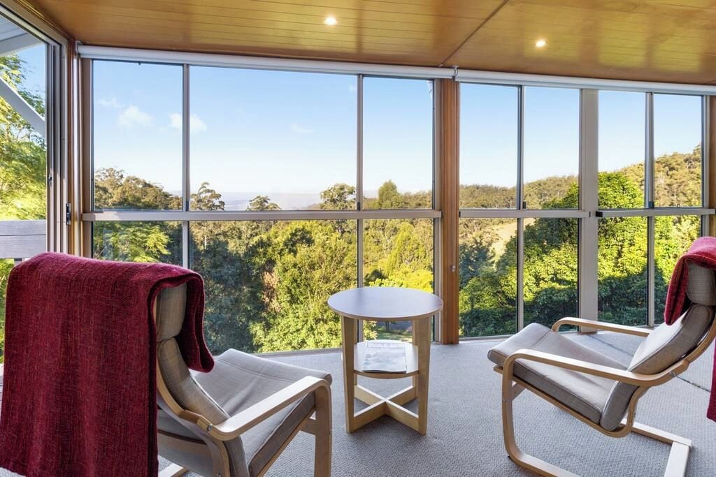 Une pièce comprenant deux fauteuils confortables, une petite table et de grandes fenêtres donnant sur un paysage boisé.