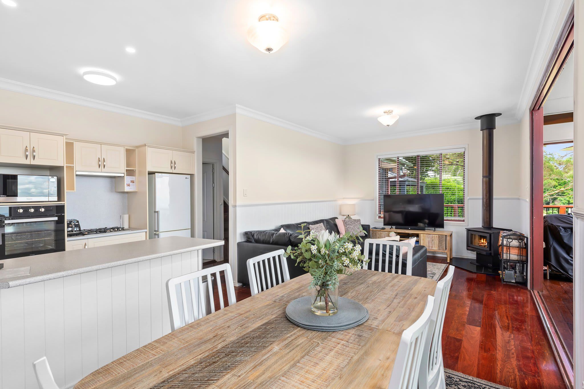 A modern kitchen with white cabinets, a dining area with a wooden table, and a living room with a sofa and TV.