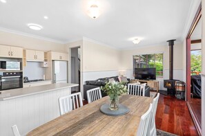 A modern kitchen with white cabinets, a dining area with a wooden table, and a living room with a sofa and TV. - Quingilli Luxury Chalet 1 Bedroom (Ravensbourne)