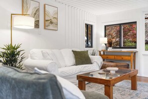 Ein modernes Wohnzimmer mit einem weißen Sofa, einem Holztisch und einem großen Fenster mit Blick auf herbstliche Laubfärbung.