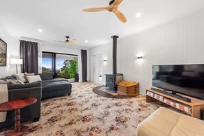 A modern living room with a sofa, a television, a fireplace, and a ceiling fan. - Mahali House - 6 Bedroom Holiday House (Ravensbourne)