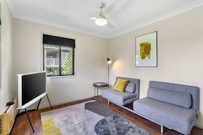 A room with a ceiling fan, a flat-screen TV on a stand, a grey sofa with a yellow pillow, a small side table with a lamp, and a framed picture on the wall. - Yume House Stylish Country Escape 1 Bedroom (Ravensbourne)