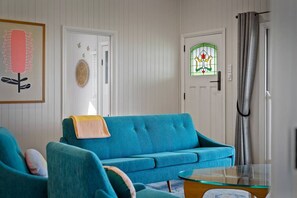 A room with a teal sofa, a glass coffee table, a door with a stained glass window, and a framed picture on the wall. - The Quince Cottage -  1 Bedroom (Ravensbourne)
