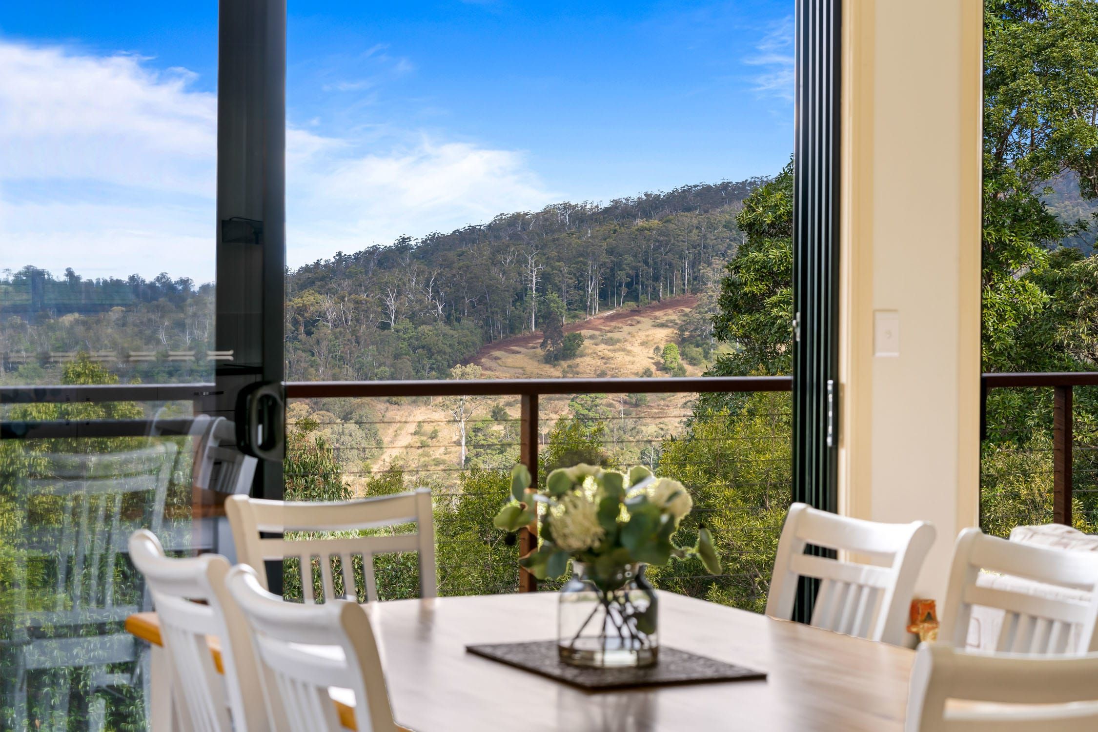 Un coin repas avec une table et des chaises, un vase rempli de fleurs et une vue sur une colline boisée.