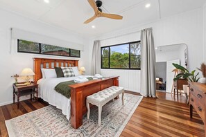 A bedroom with a wooden bed, a bench, a nightstand, a dresser, a ceiling fan, and a large window with a view. - Mahali House - 5 Bedroom Holiday House (Ravensbourne)