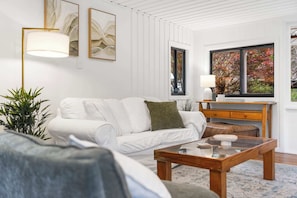 A modern living room with a white sofa, a wooden coffee table, and a large window with autumn foliage view. - Mahali House - 5 Bedroom Holiday House (Ravensbourne)