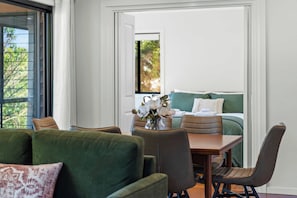 Une chambre moderne avec un canapé vert, une table en bois et un lit doté d’une tête de lit verte. Une fenêtre offre une vue sur la verdure à l’extérieur.