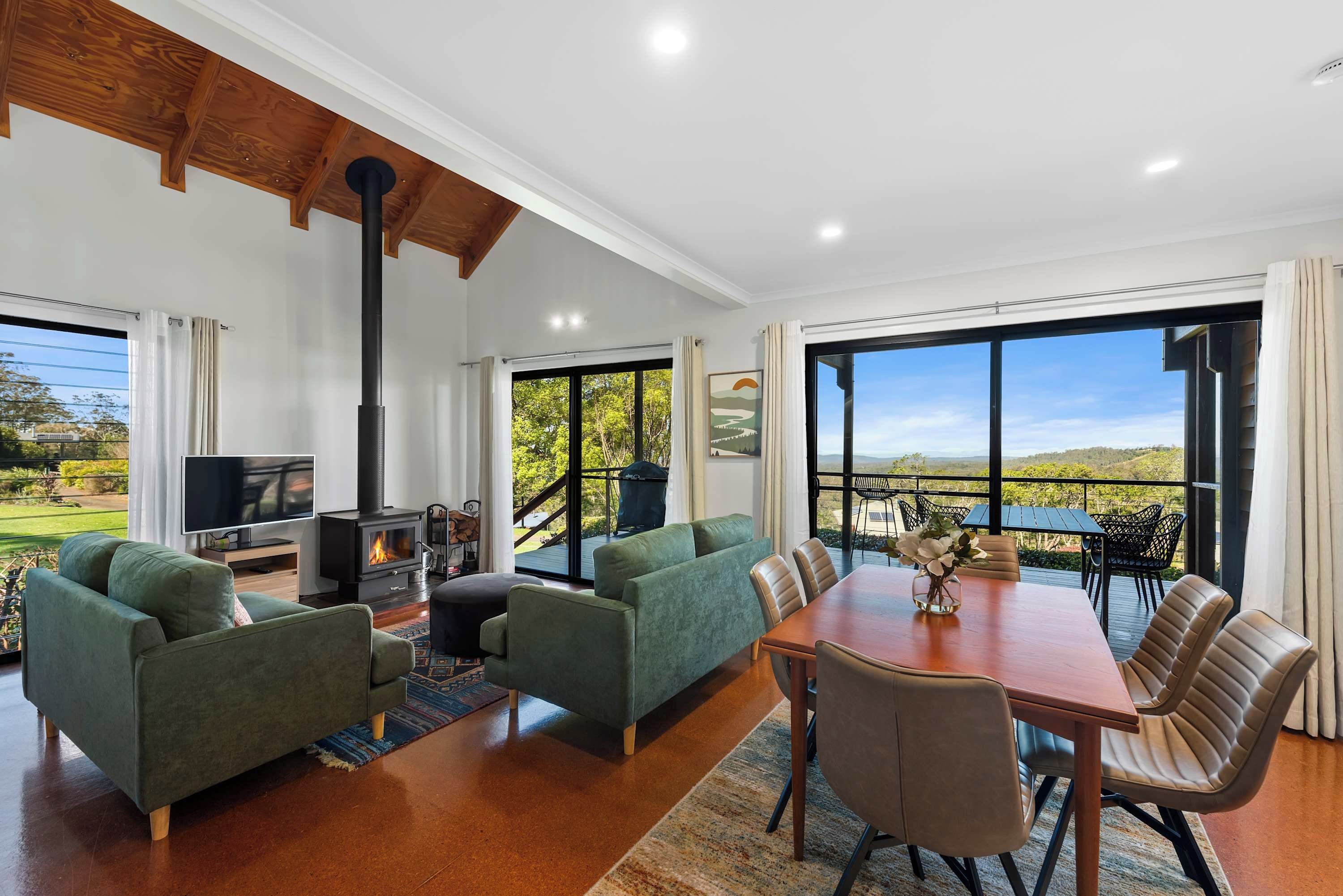 A spacious living room with a fireplace, green sofas, a wooden dining table with chairs, and large windows offering a view of the outdoors.