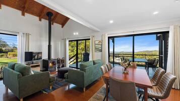 A spacious living room with a fireplace, green sofas, a wooden dining table with chairs, and large windows offering a view of the outdoors.