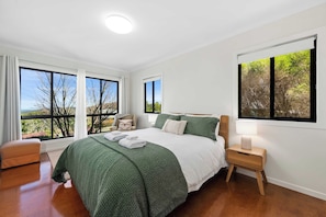 A bedroom with a large bed, a nightstand, a chair, and a view of trees through the windows. - Havenwood Chalet 1 Bedroom (Ravensbourne)