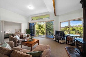 A spacious living room with a large TV, comfortable seating, a fireplace, and a balcony with a view of trees. - Kookaburra Kottage Luxury Chalet 1 Bedroom (Ravensbourne)