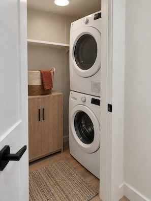 A laundry room with two stacked washing machines, a wooden cabinet, and a woven basket with a towel. - NYC Portal , 3BD/2BA , Parking & Laundry , 20m NYC (Newark)