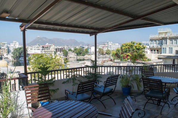 A covered outdoor seating area with multiple tables and benches, overlooking a cityscape with buildings and a mountain in the distance. - Hotel Lake Ghat Palace (Udaipur)