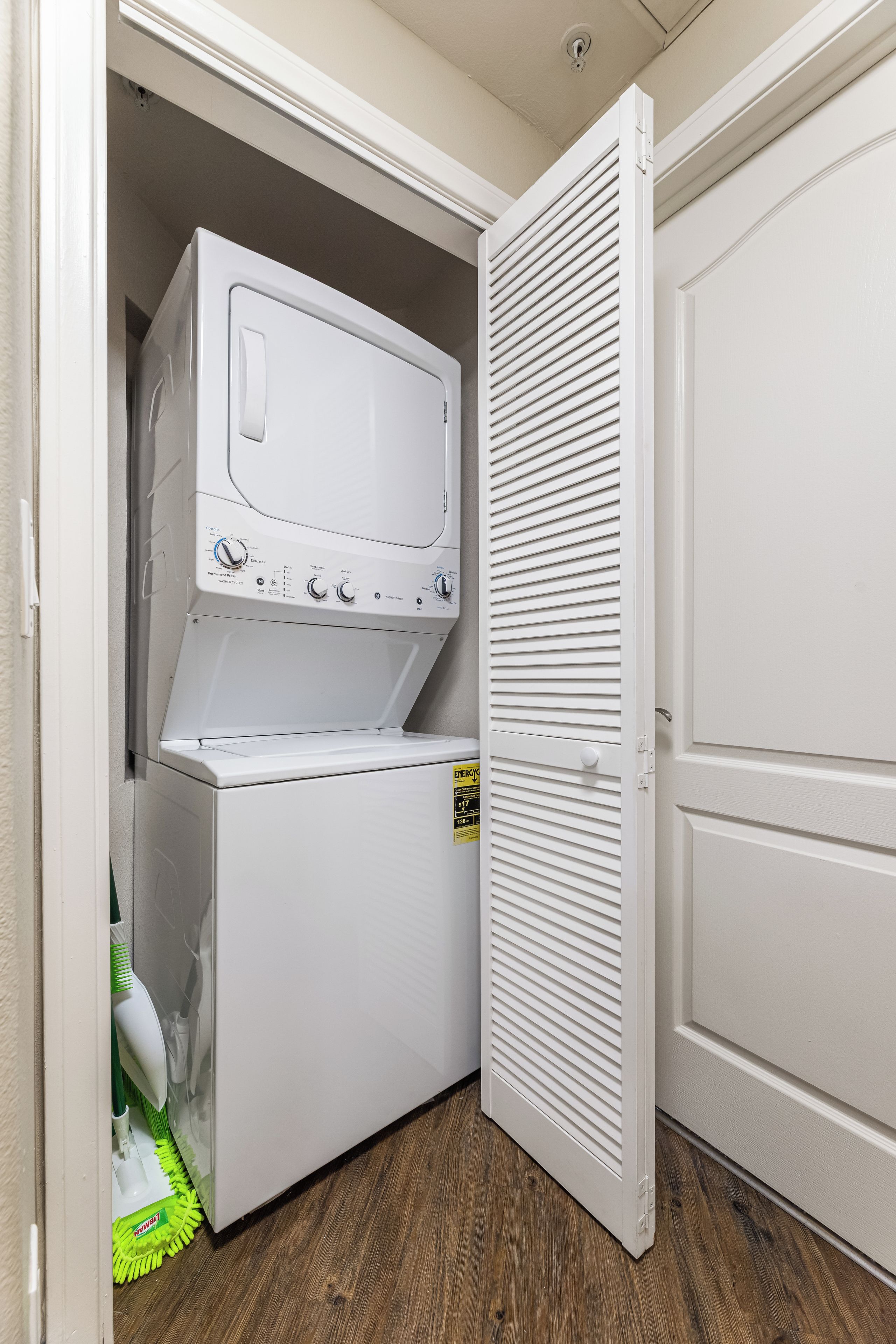 A laundry room with a front-loading washer and dryer, a door with horizontal slats, and a wooden floor.