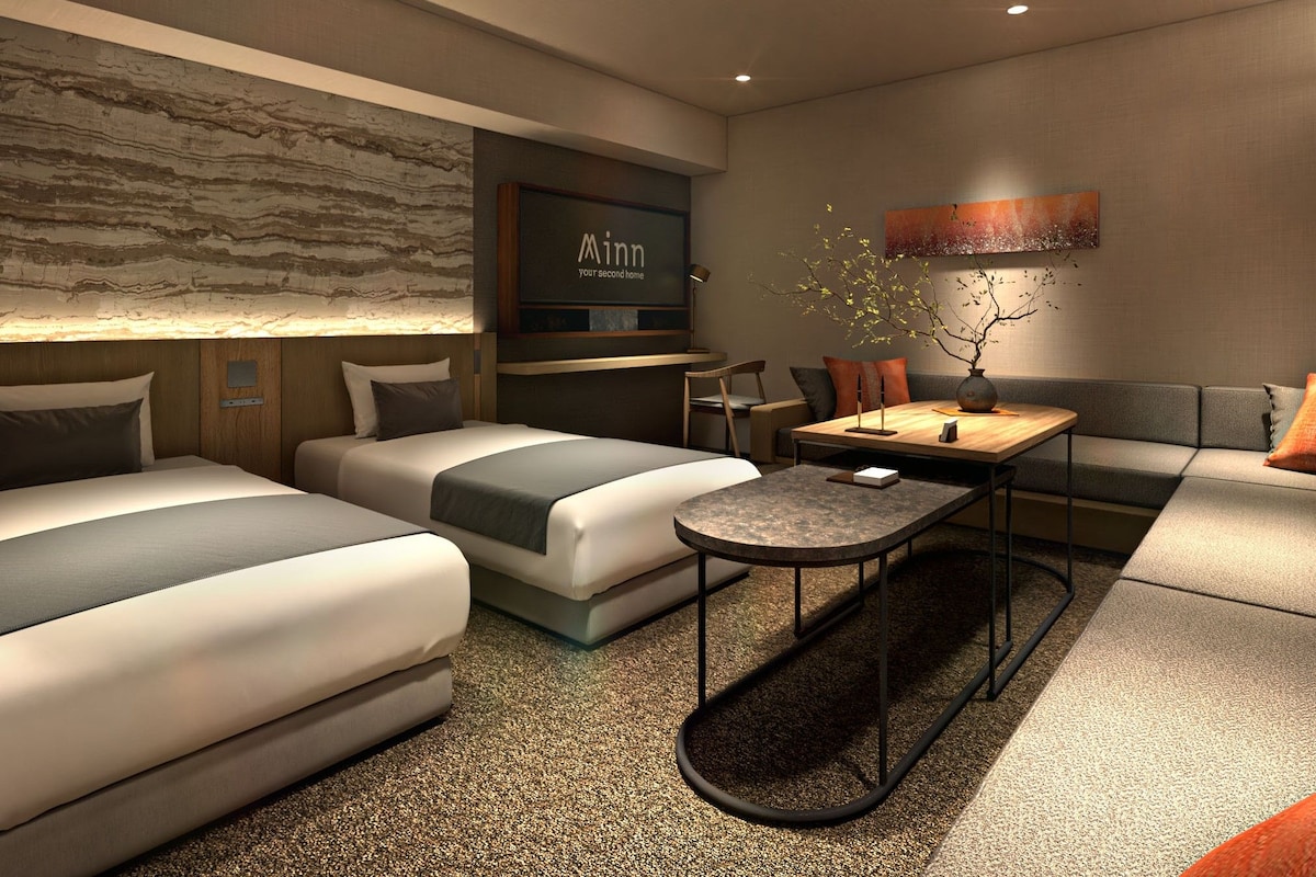 A modern hotel room with two beds, a small table, a sofa, and a decorative wall piece.