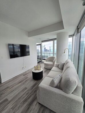 A modern living room with a flat-screen TV, a round coffee table with decorative items, a beige sofa, and a view of the city through large windows. - Toronto in the Sky | CN Tower & Lake Views (Toronto)