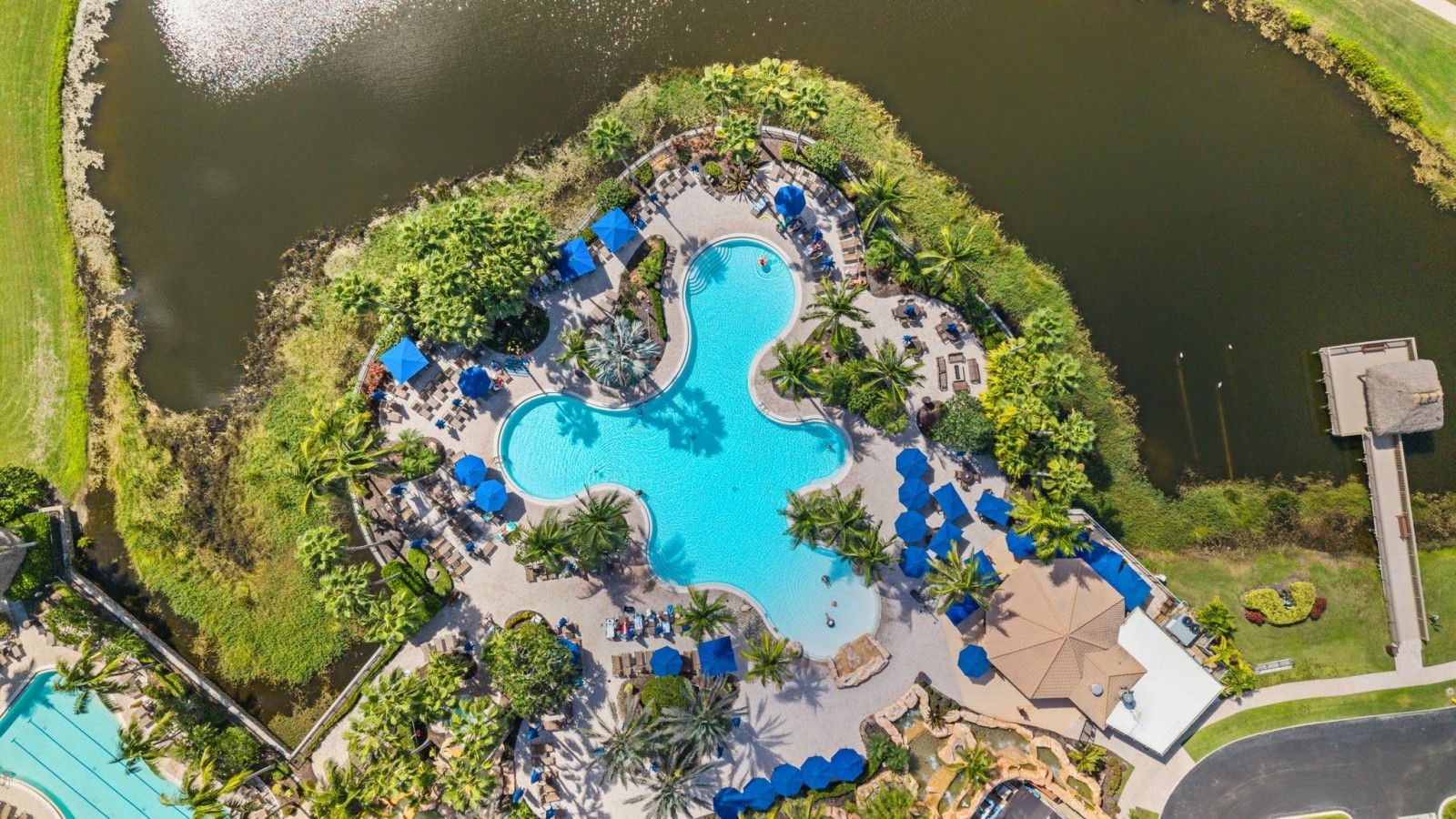 An aerial view of a resort with a central swimming pool, surrounded by lounge areas and greenery.