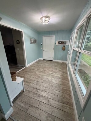 A hallway with a wooden floor, a white door, and a window with a view of greenery. - Sand Lake Extended Stay Ranch - Fully Furnished Lake Home - Monthly Stays (Lindenhurst)