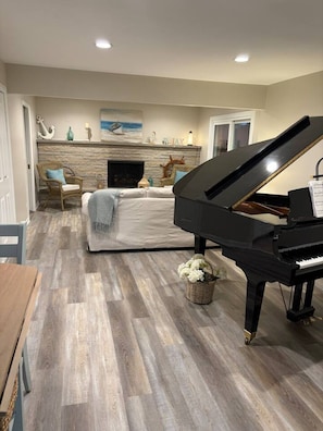 A living room with a grand piano, a sofa, a fireplace, and a stone wall. - Sand Lake Extended Stay Ranch - Fully Furnished Lake Home - Monthly Stays (Lindenhurst)