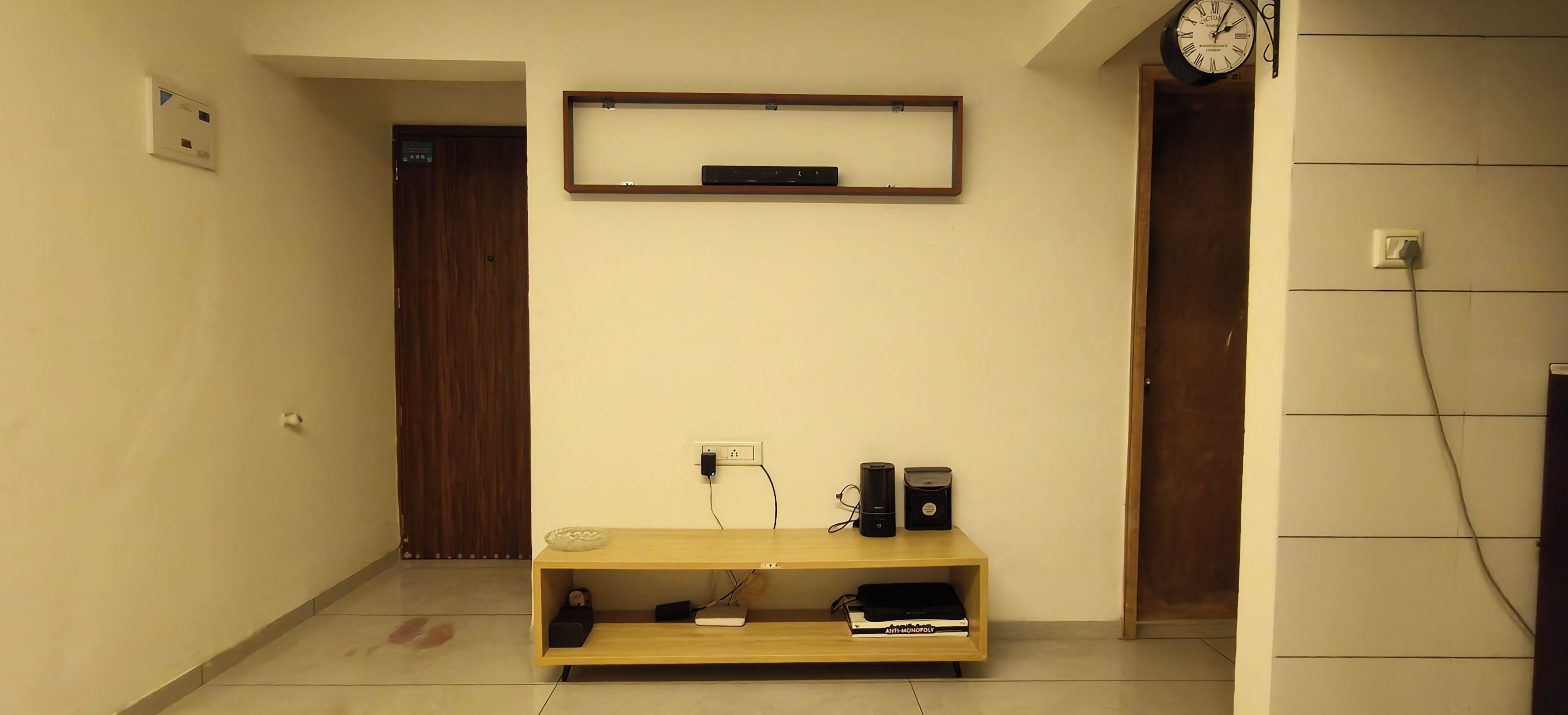 Un couloir avec une porte en bois, une horloge et une étagère contenant des livres et des appareils électroniques.