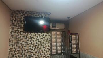 A room with a patterned wall, a flat-screen TV mounted above a window, and an air conditioning unit.