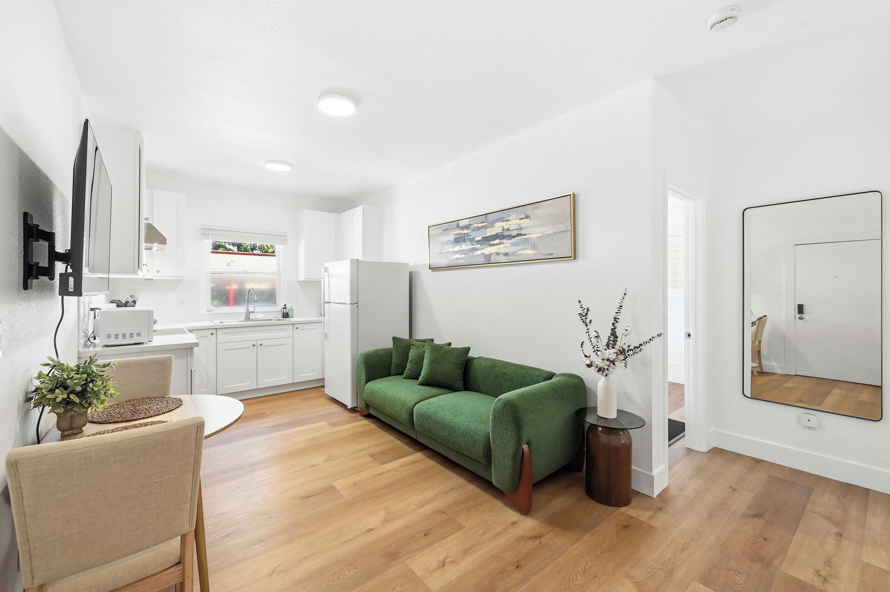 A modern living room with a green sofa, a dining area with a table and chairs, a kitchen with white cabinets, and a large mirror on the wall.