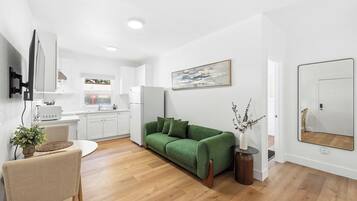 A modern living room with a green sofa, a dining area with a table and chairs, a kitchen with white cabinets, and a large mirror on the wall.