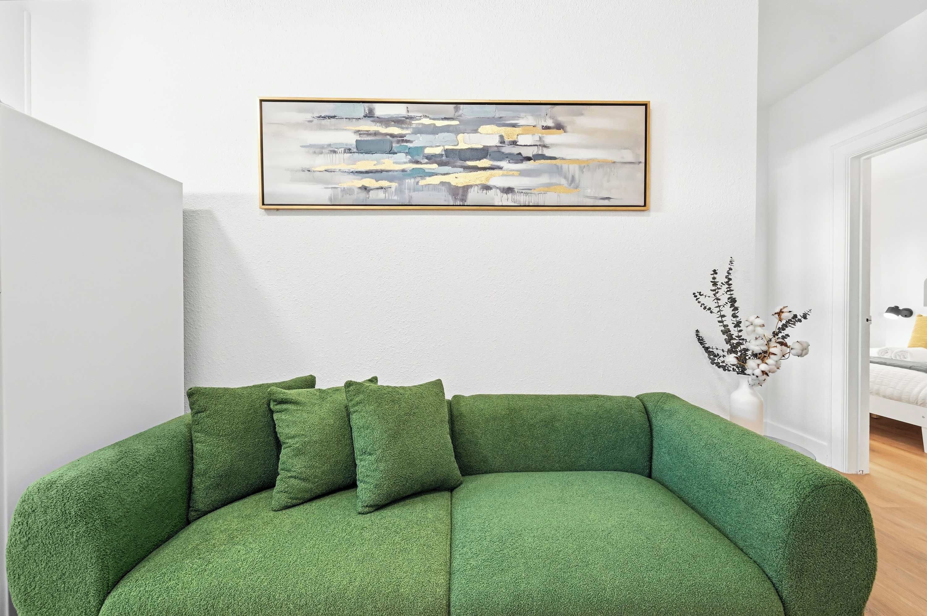 A green sofa with cushions, a wall-mounted abstract painting, and a vase with branches and cotton.
