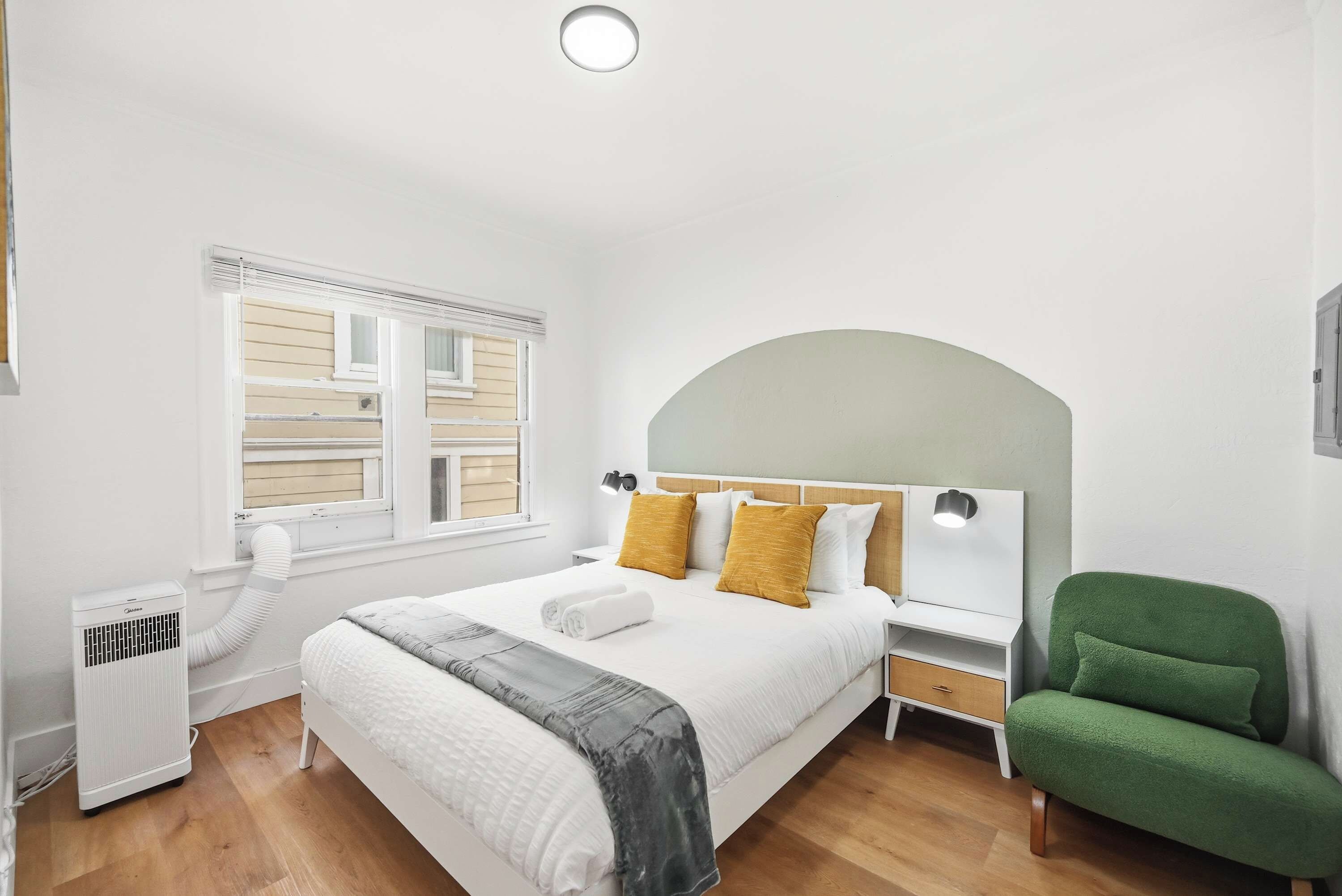 A hotel room with a bed, a nightstand, a green chair, a window with shutters, and a radiator.