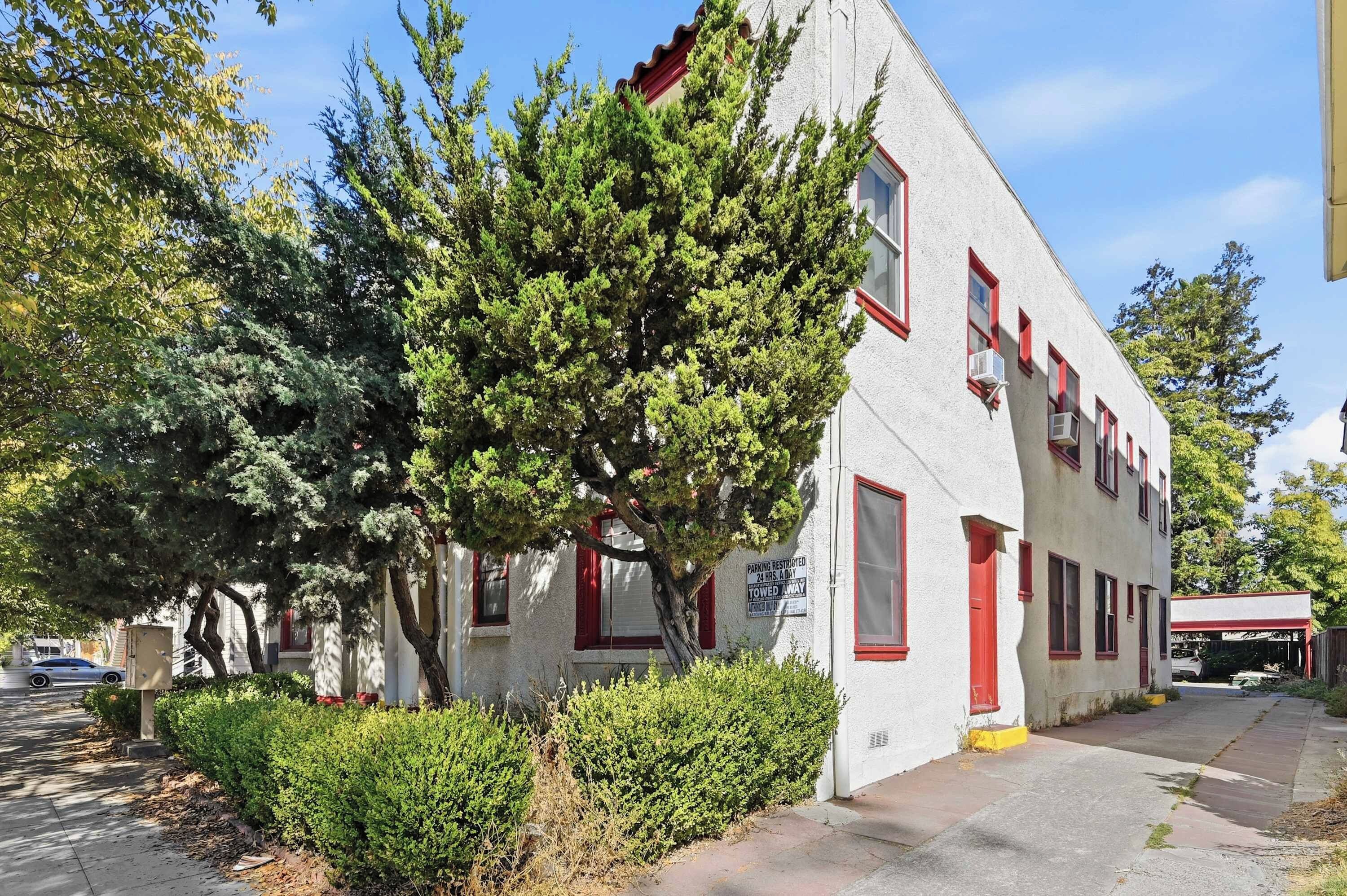 A white building with red trim and multiple windows, surrounded by trees and a sidewalk.