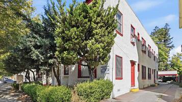 A white building with red trim and multiple windows, surrounded by trees and a sidewalk.
