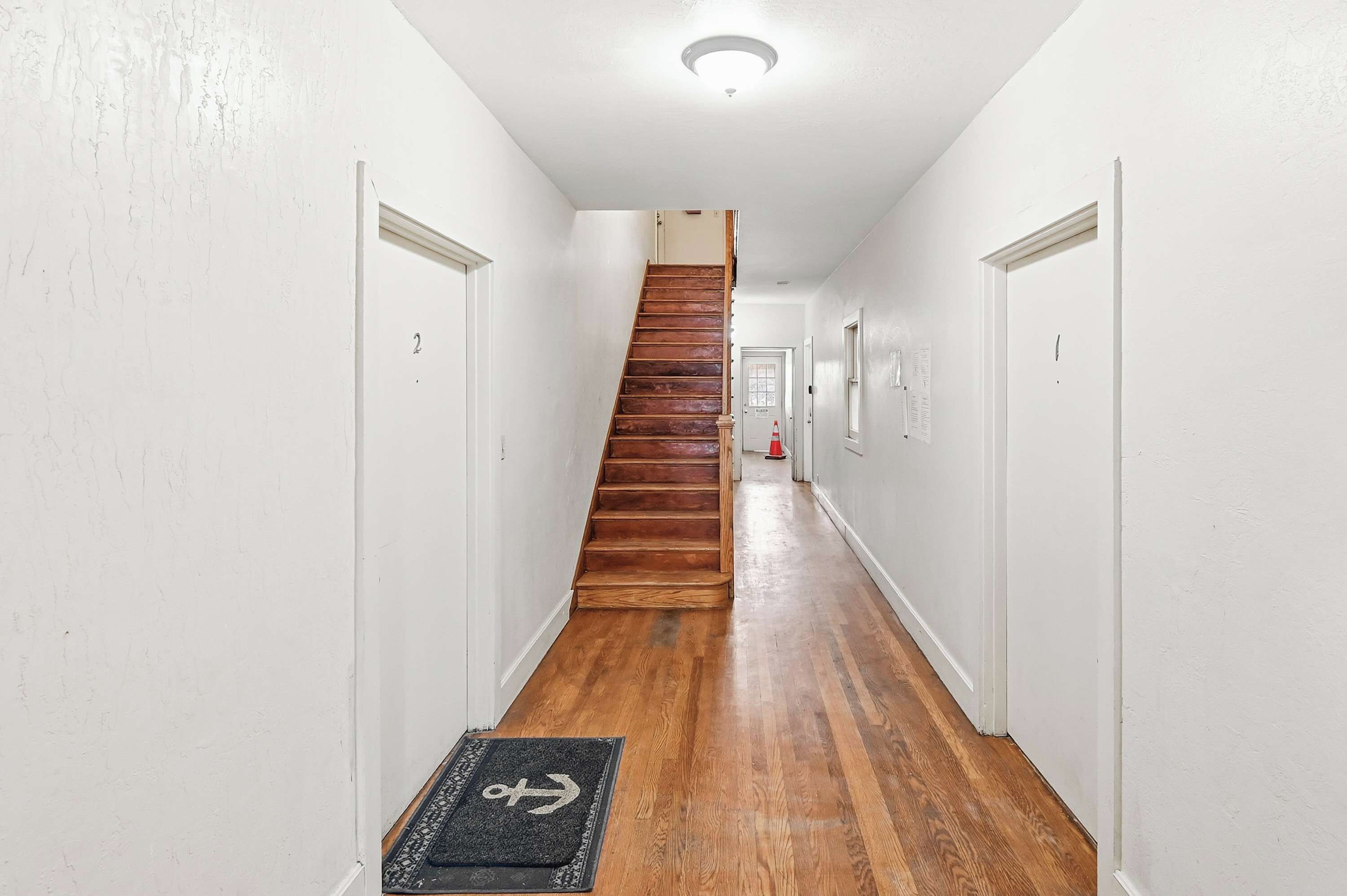 A narrow hallway with wooden flooring, white walls, and a wooden staircase leading to an upper level.
