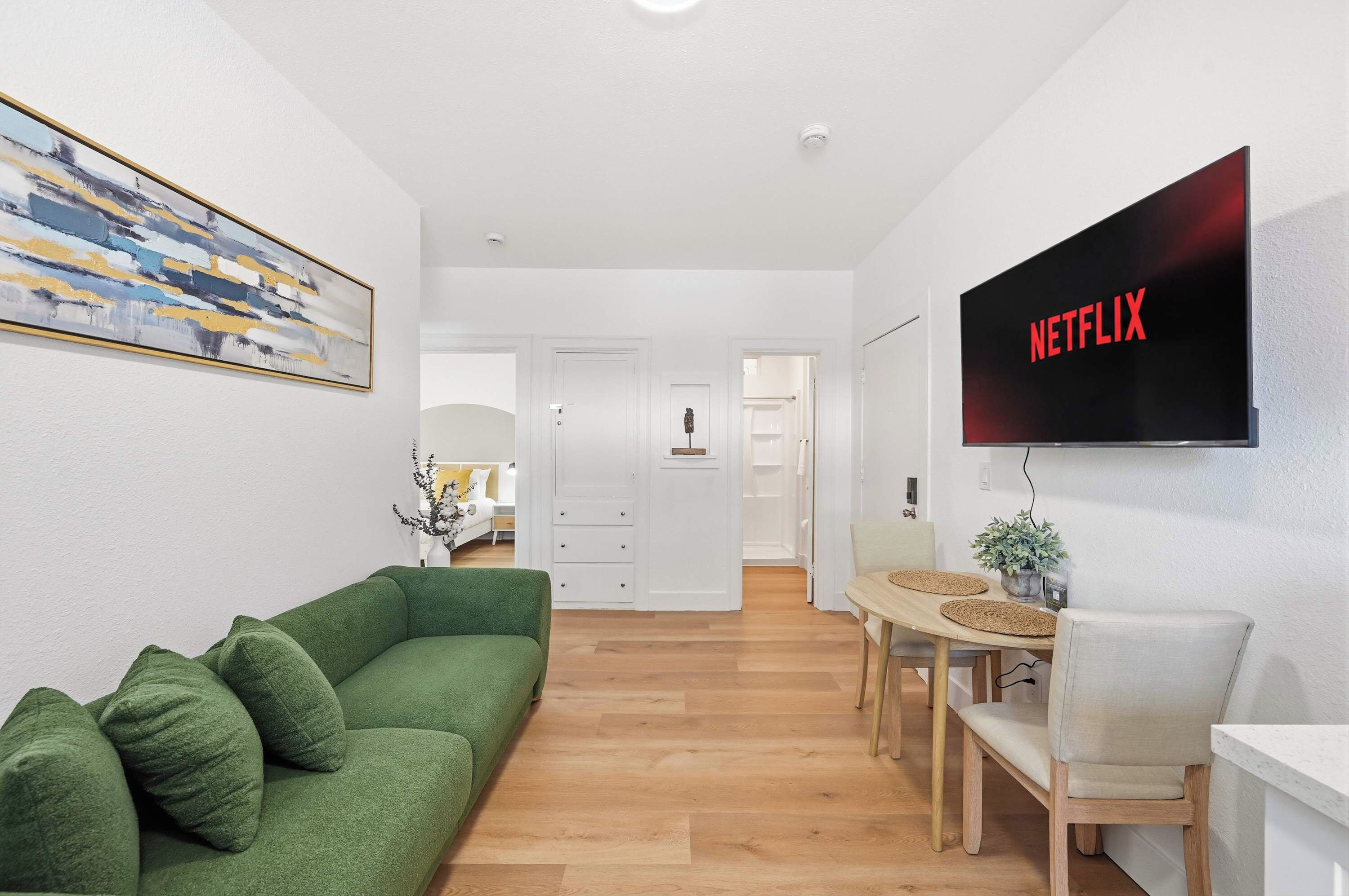 A modern living room with a green sofa, a dining area with a wooden table, and a flat-screen TV mounted on the wall displaying 'NETFLIX'.